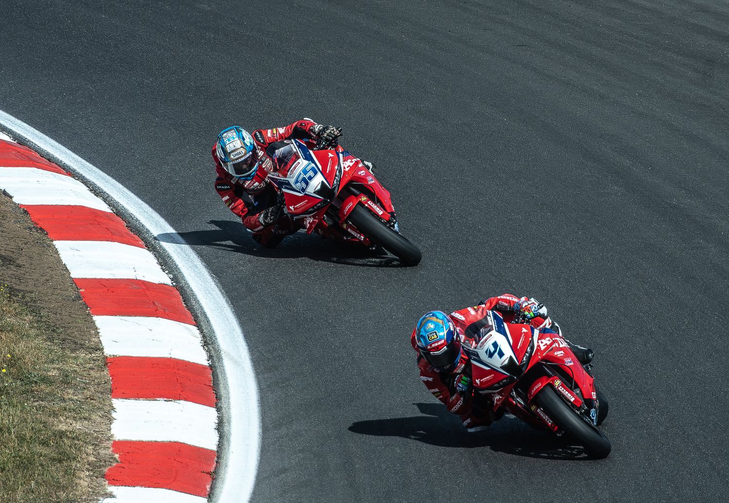 Two motorcycle racers leaning into a corner past a red and white kerb, motorsport photography