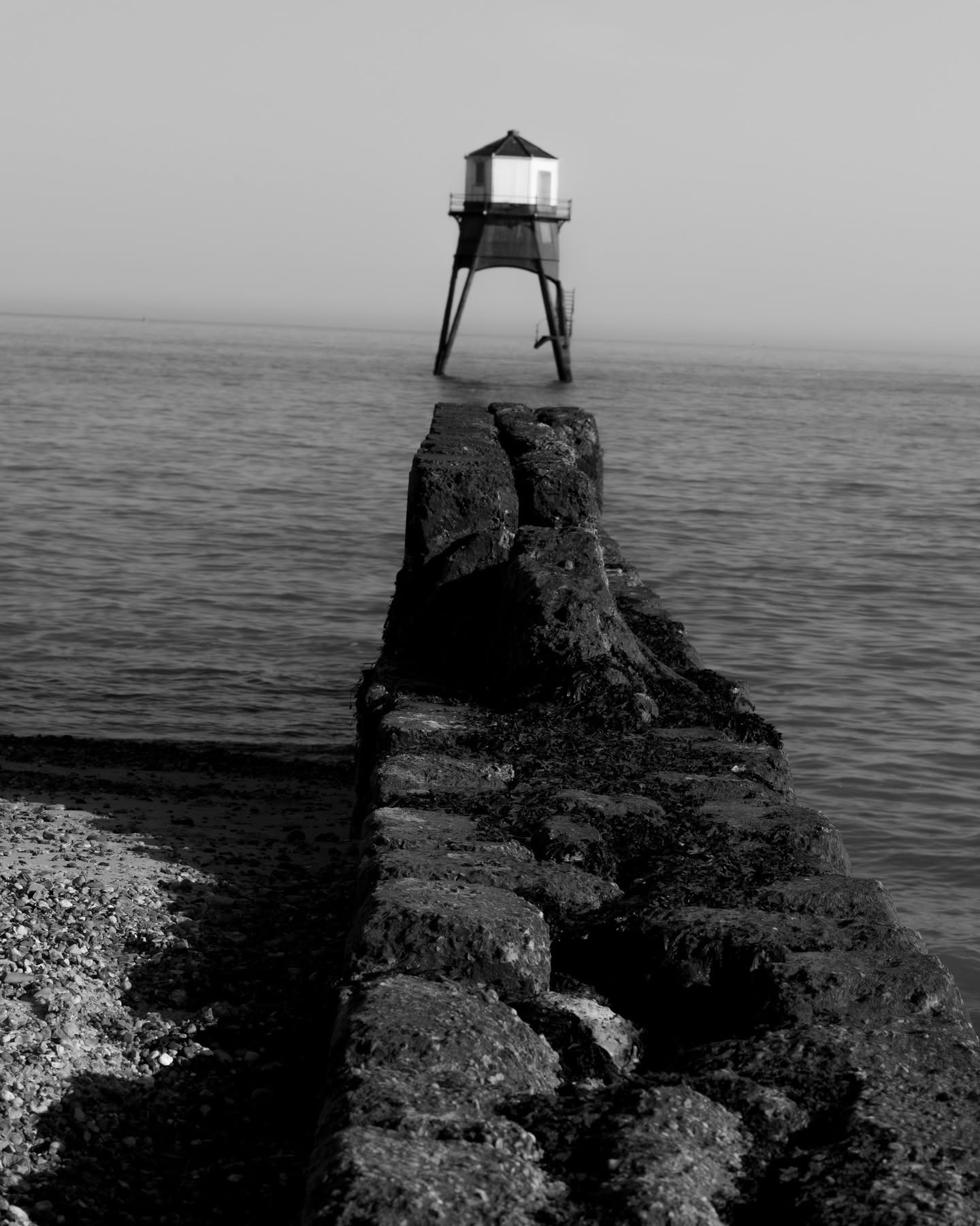 Wooden breakwater leading out to a lighthouse at sea, black and white Suffolk coast landscape photography