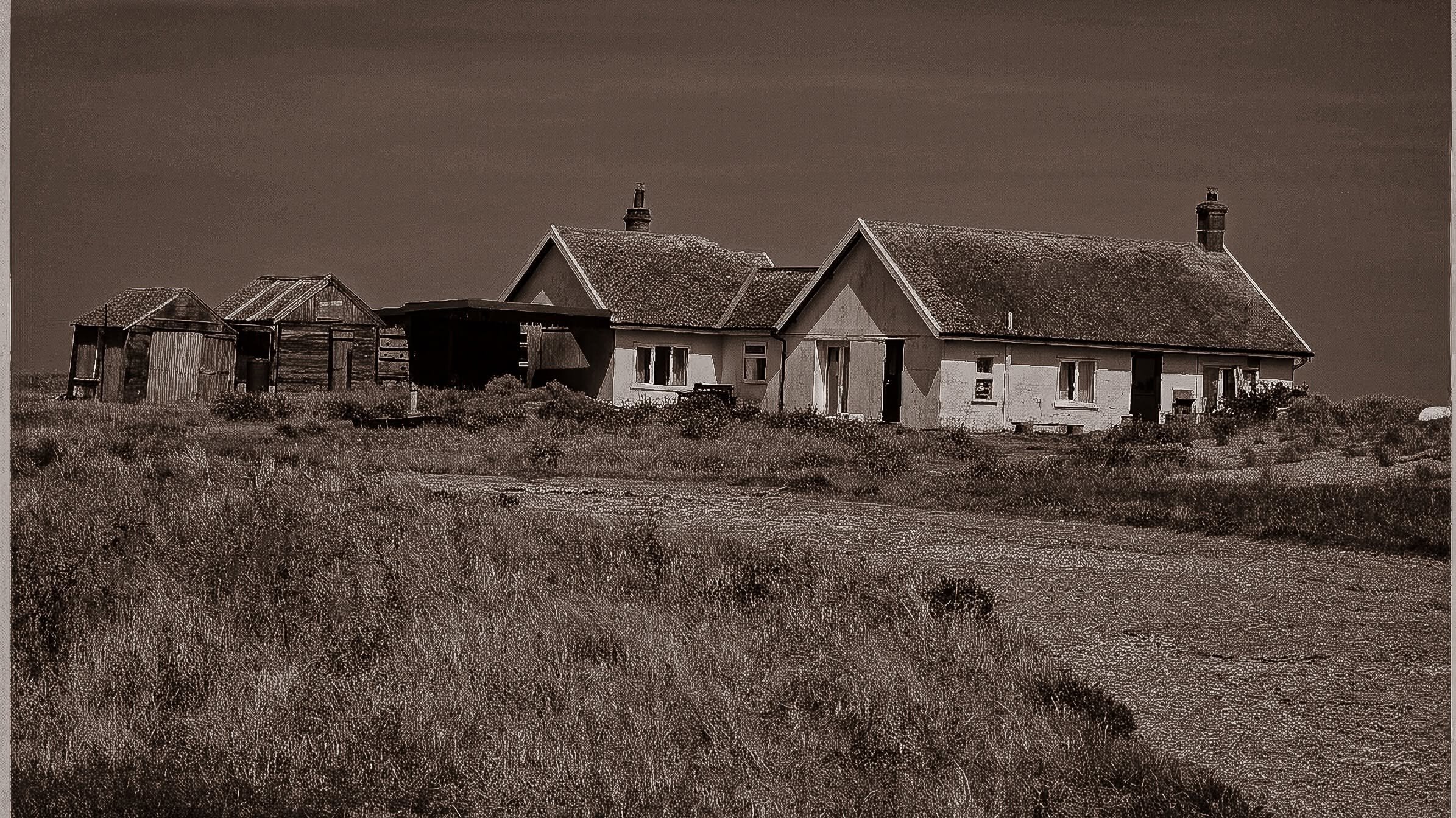 Row of abandoned coastal cottages and sheds in long grass, sepia-toned architecture photography