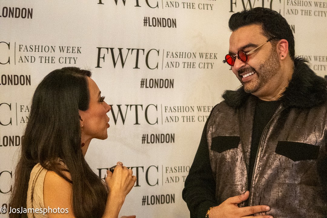 Samira Ebrahimi chatting with a guest in red-tinted glasses at the Fashion Week and the City press wall, London Fashion Week 2026